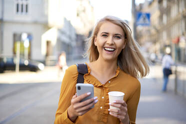 Porträt einer glücklichen jungen Frau mit Smartphone und Kaffee zum Mitnehmen in der Stadt - BSZF01345