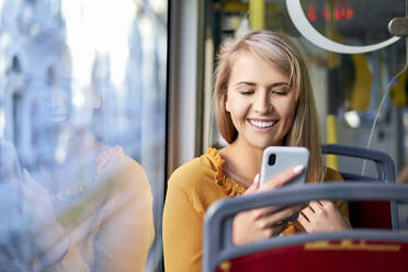 Lächelnde junge Frau, die in einer Straßenbahn ihr Smartphone benutzt - BSZF01346