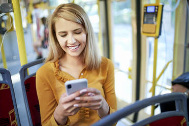 Lächelnde junge Frau, die in einer Straßenbahn ihr Smartphone benutzt - BSZF01349