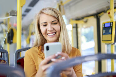 Lächelnde junge Frau, die in einer Straßenbahn ihr Smartphone benutzt - BSZF01350