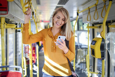 Lächelnde junge Frau, die in einer Straßenbahn ihr Smartphone benutzt - BSZF01351