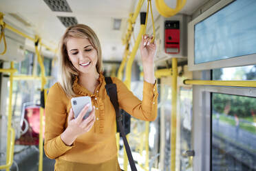 Lächelnde junge Frau, die in einer Straßenbahn ihr Smartphone benutzt - BSZF01353
