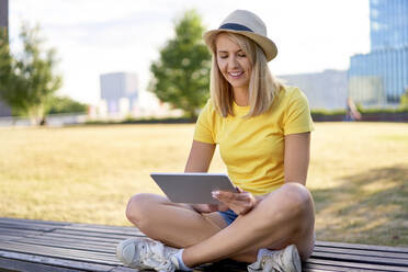 Lächelnde junge Frau sitzt auf einer Bank in der Stadt und benutzt ein Tablet - BSZF01376