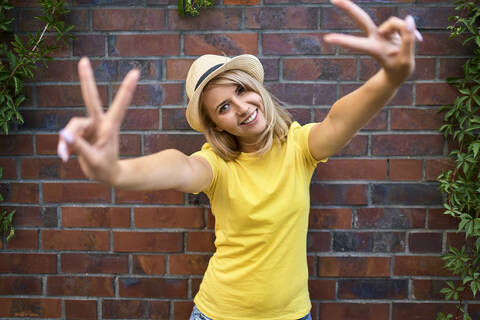 Portrait of happy young woman standing at a brick wall making peace sign stock photo