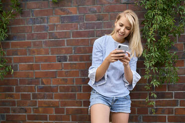 Smiling young woman standing at a brick wall using smartphone - BSZF01384