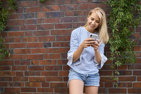 Lächelnde junge Frau, die an einer Mauer steht und ein Smartphone benutzt - BSZF01384