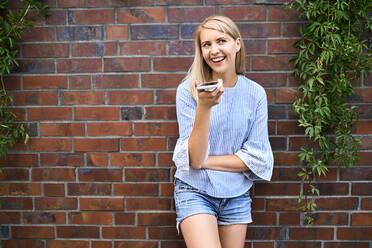 Happy young woman standing at a brick wall using smartphone - BSZF01386