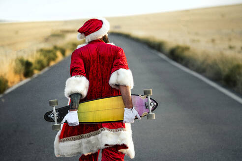 Rückansicht des Weihnachtsmanns mit einem Longboard auf der Landstraße - JCMF00179