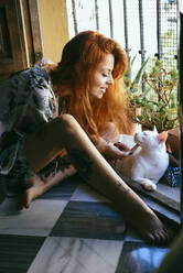 Red-haired tattooed woman playing with her cat at home - JSMF01240