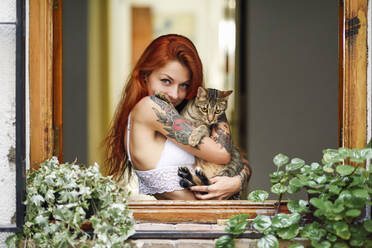 Portrait of red-haired tattooed woman with her cat at the window - JSMF01254