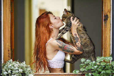 Red-haired tattooed woman with her cat at the window - JSMF01255