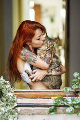 Red-haired tattooed woman with her cat at the window - JSMF01256