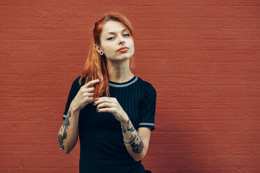 Portrait of red-haired tattooed woman standing at a brick wall - JSMF01263