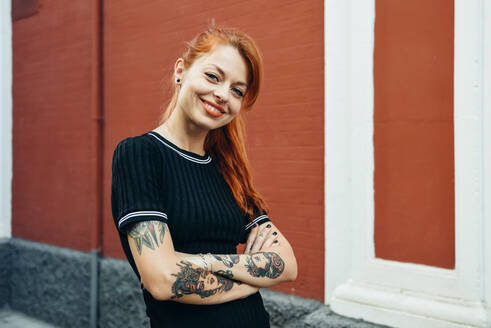 Portrait of smiling red-haired tattooed woman standing in the city - JSMF01264
