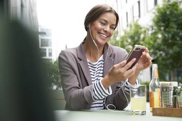 Happy businesswoman with earphones listening to music at an outdoors cafe - PNEF01859