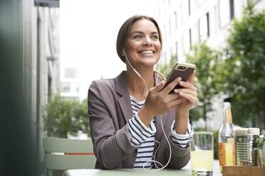 Happy businesswoman with earphones listening to music at an outdoors cafe - PNEF01860