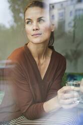 Porträt einer nachdenklichen Frau mit Getränk hinter einer Fensterscheibe - PNEF01907