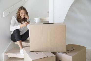 Woman sitting on staircase with smart phone and cardboard boxes - FOLF10460