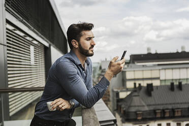 Junger Mann schaut auf dem Balkon auf sein Handy - FOLF10520