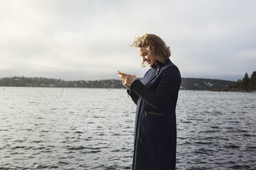 Junge Frau steht neben einem See und hält ein Handy in der Hand - FOLF10651