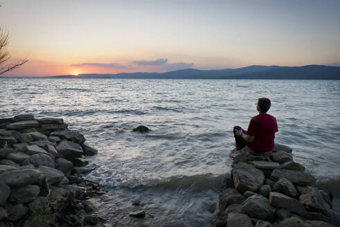 Rückansicht eines reifen Mannes, der bei Sonnenuntergang auf einem Felsen sitzend den Trasimenischen See betrachtet, Isola Maggiore, Italien - LOMF00907