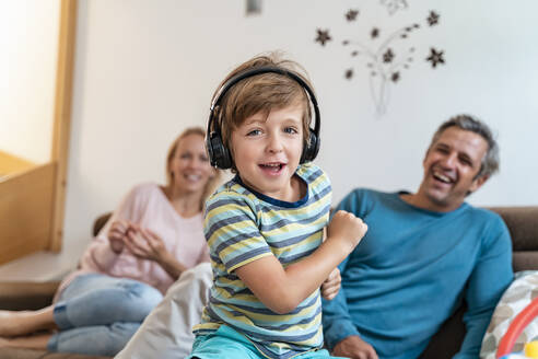 Porträt eines Jungen, der zu Hause auf der Couch mit Kopfhörern Musik hört, mit den Eltern im Hintergrund - DIGF08201