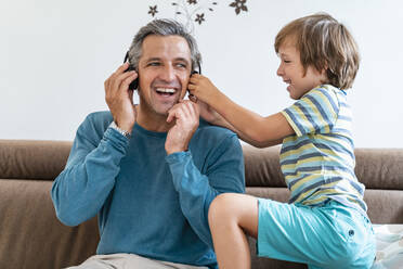 Happy father with boy on couch at home listening to music with headphones - DIGF08204