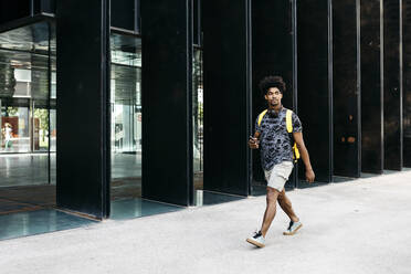 Mann mit gelbem Rucksack, Smartphone und Kopfhörern geht die Straße entlang, Barcelona, Spanien - JRFF03687