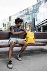 Man sitting on the bench listening music with headphones while using mobile phone, Barcelona, Spain - JRFF03697