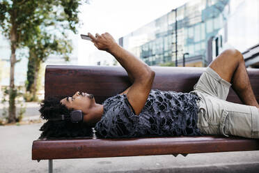 Man lying on bench listening music with headphones while using mobile phone, Barcelona, Spain - JRFF03701