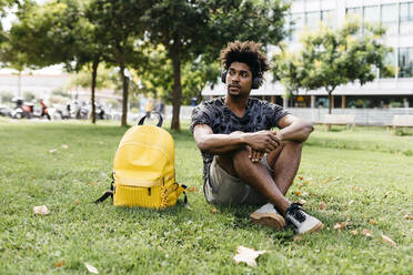 Portrait of man sitting on a meadow listening music with cordless headphones, Barcelona, Spain - JRFF03704