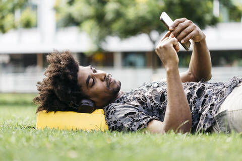 Mann liegt auf einer Wiese und hört mit Kopfhörern Musik, während er ein Mobiltelefon benutzt, Barcelona, Spanien, lizenzfreies Stockfoto