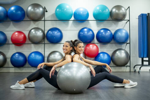 Portrait of smiling twin sisters sitting on the floor in gym with a fitness ball - OCMF00656