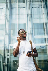Glücklicher junger Mann mit Mobiltelefon in der Stadt, der einen Kopfhörer einsteckt - OCMF00685