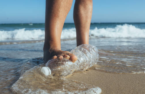 Fuß eines Mannes, der auf eine am Strand liegende leere Plastikflasche tritt, Nahaufnahme - CJMF00005