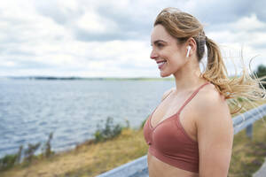 Porträt einer lächelnden sportlichen Frau mit drahtlosen Kopfhörern am Seeufer - BSZF01388