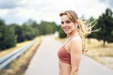 Porträt einer lächelnden sportlichen Frau mit drahtlosen Kopfhörern in der Natur - BSZF01403