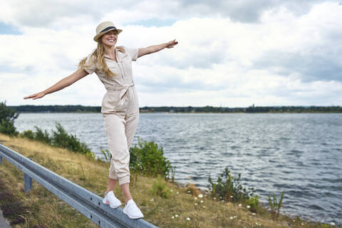 Glückliche Frau balanciert auf der Leitplanke am Seeufer, lizenzfreies Stockfoto