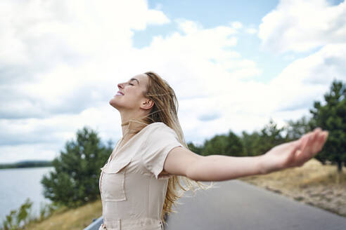 Happy woman standing at the lakeside with outstretched arms - BSZF01418