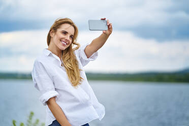 Glückliche Frau nimmt Selfie mit Smartphone am Seeufer - BSZF01443