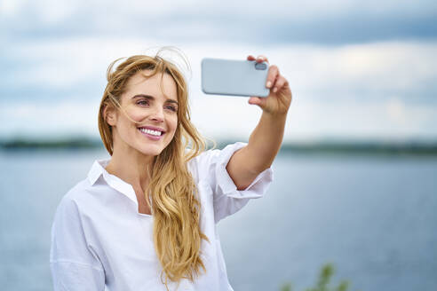 Glückliche Frau nimmt Selfie mit Smartphone am Seeufer - BSZF01444