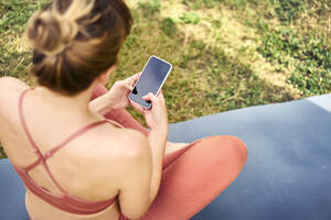 Frau, die ihr Smartphone benutzt, während sie eine Pause von der Yoga-Praxis im Freien macht - BSZF01453