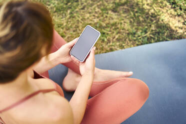 Frau, die ihr Smartphone benutzt, während sie eine Pause von der Yoga-Praxis im Freien macht - BSZF01454