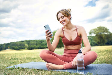 Lächelnde Frau, die ihr Smartphone benutzt, während sie eine Pause vom Yoga im Freien macht - BSZF01461