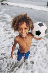 Porträt eines lächelnden Jungen, der einen Fußball am Strand hält - LJF00978