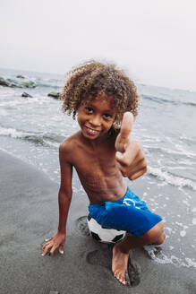 Lächelnder Junge, der am Strand sitzt und die Daumen nach oben zeigt - LJF00989