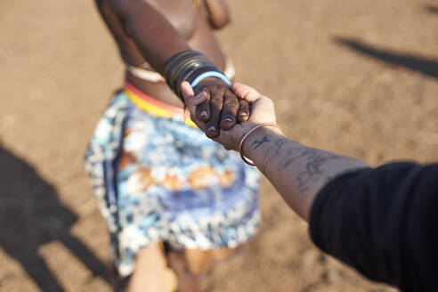 Traditionelle Muhacaona-Frau und weiße Frau halten sich an den Händen, Oncocua, Angola - VEGF00640