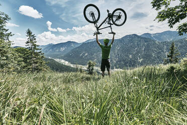 Mountainbiker in den Bergen, der sein Mountainbike hochhält, Bayern, Deutschland - WFF00089