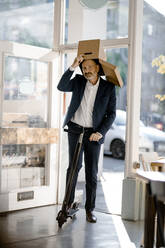 Businessman with cardboard box on his head, riding e-scooter in a coffee shop - KNSF06373