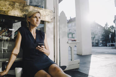 Businesswoman in coffee shop, sitting on ground, using smartphone - KNSF06378